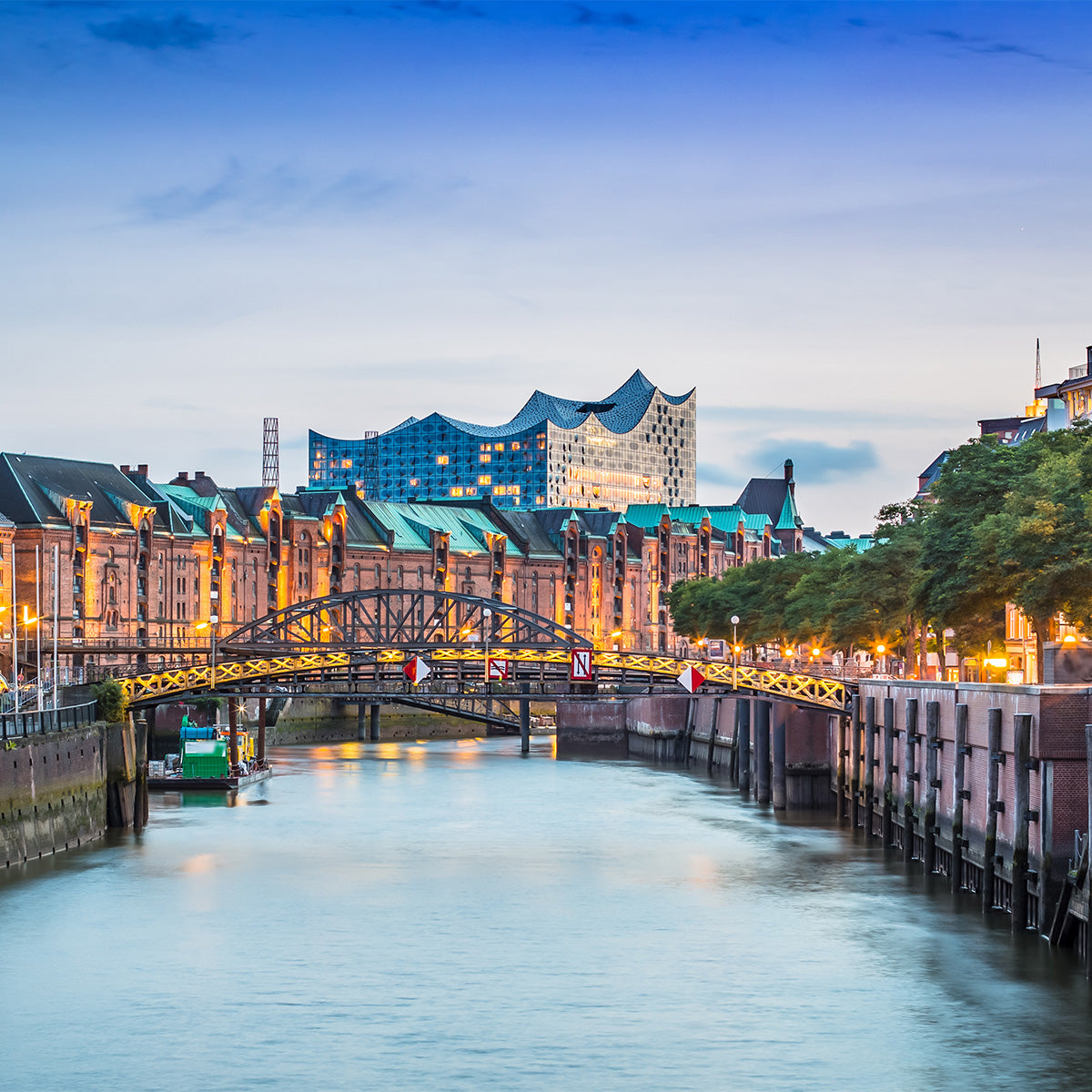 Evening view of Hamburg's Speicherstadt with historic red-brick warehouses, a canal with a small bridge in the foreground, and the illuminated Elbphilharmonie concert hall in the background
