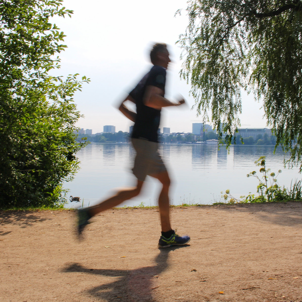 Blurred motion of a person jogging along a lakeside path on a sunny day, with trees framing the view and a city skyline visible across the water.