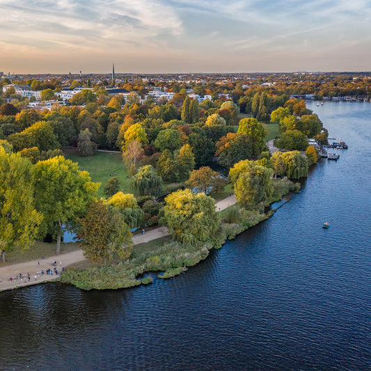 Elevated view of a large lakeside park with autumn-colored trees, walking paths, and people strolling along the shore, with a city skyline and church spire in the distance.